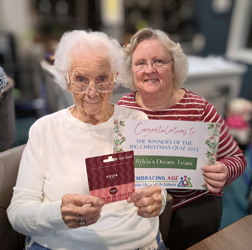 Sylvia, a female care home resident, and her daughter Lynette sit smiling. Sylvia is holding their Costa Coffee prize voucher and Lynette is holding a certificate congratulating Sylvia's Dream Team on winning the Big Christmas Quiz 2023